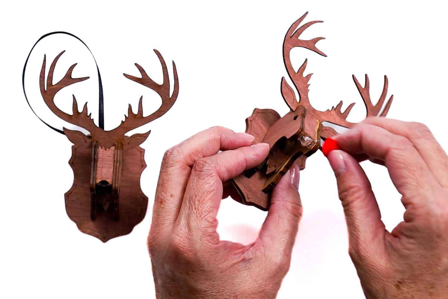 Hands attaching a small red pom-pom nose to a finished wooden reindeer ornament, with another completed reindeer ornament visible in the background.
