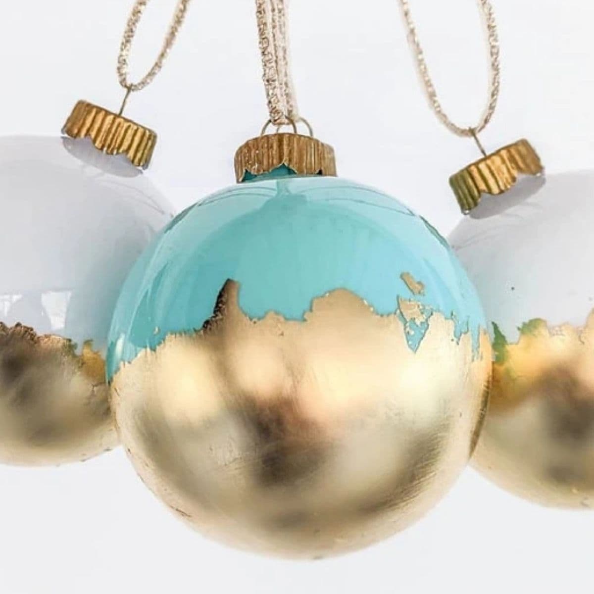 Three hanging Christmas ornaments with gold leaf bottoms, including one teal and two white designs, shown close up against a light background.