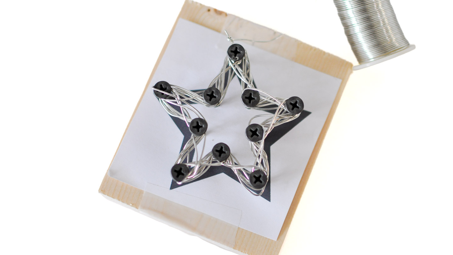 A star-shaped wire ornament being formed by wrapping silver wire around screws placed on a star template attached to a small wood block.
