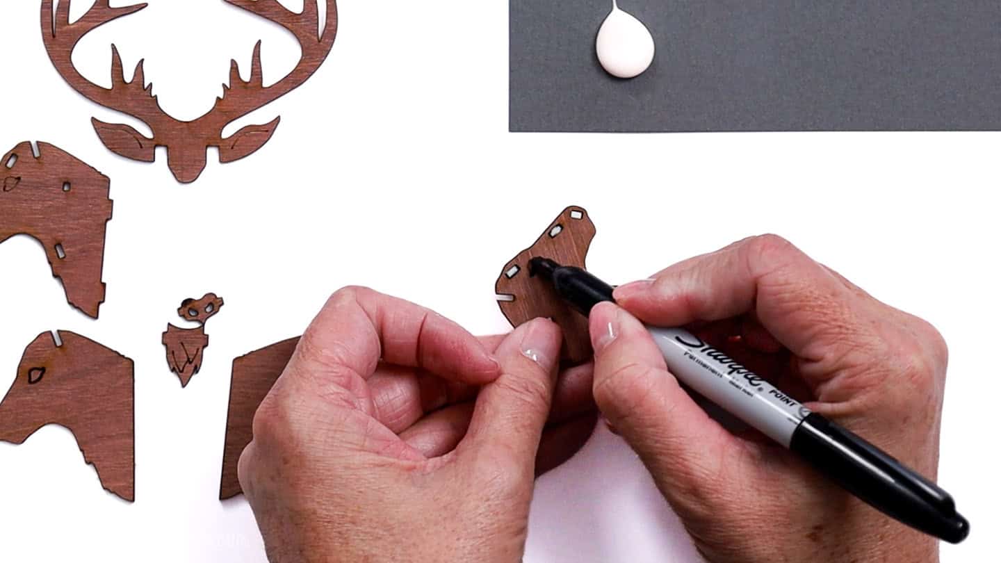 Hands using a black Sharpie to add details to a small laser-cut wooden reindeer piece, with other reindeer ornament parts laid out on the table.
