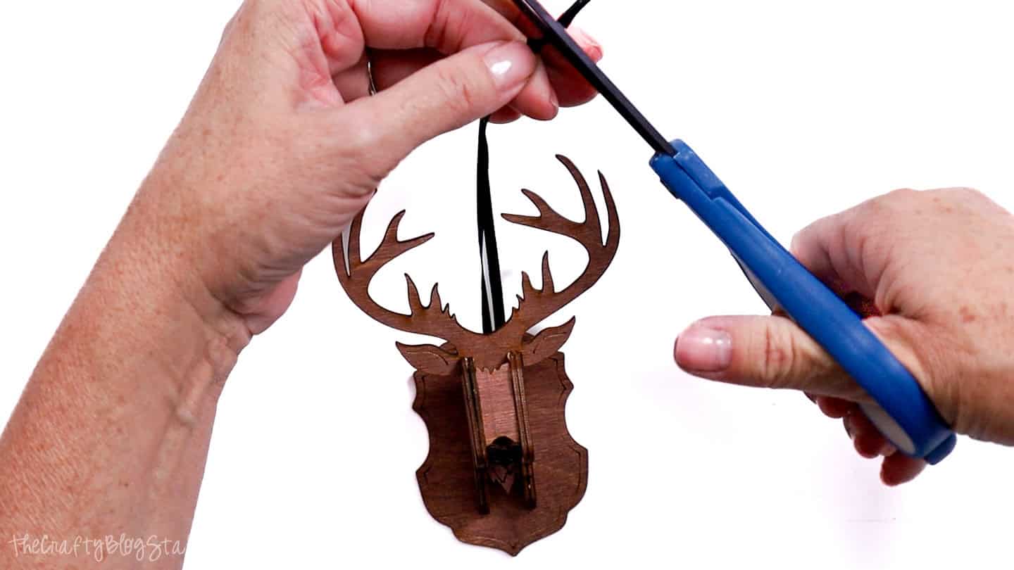 Hands trimming a black ribbon hanger with scissors on a finished wooden reindeer ornament with antlers and plaque.