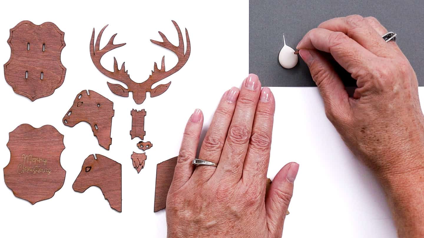 Hands dipping a small laser-cut wooden piece into a drop of wood glue, with other reindeer ornament pieces laid out on the table.