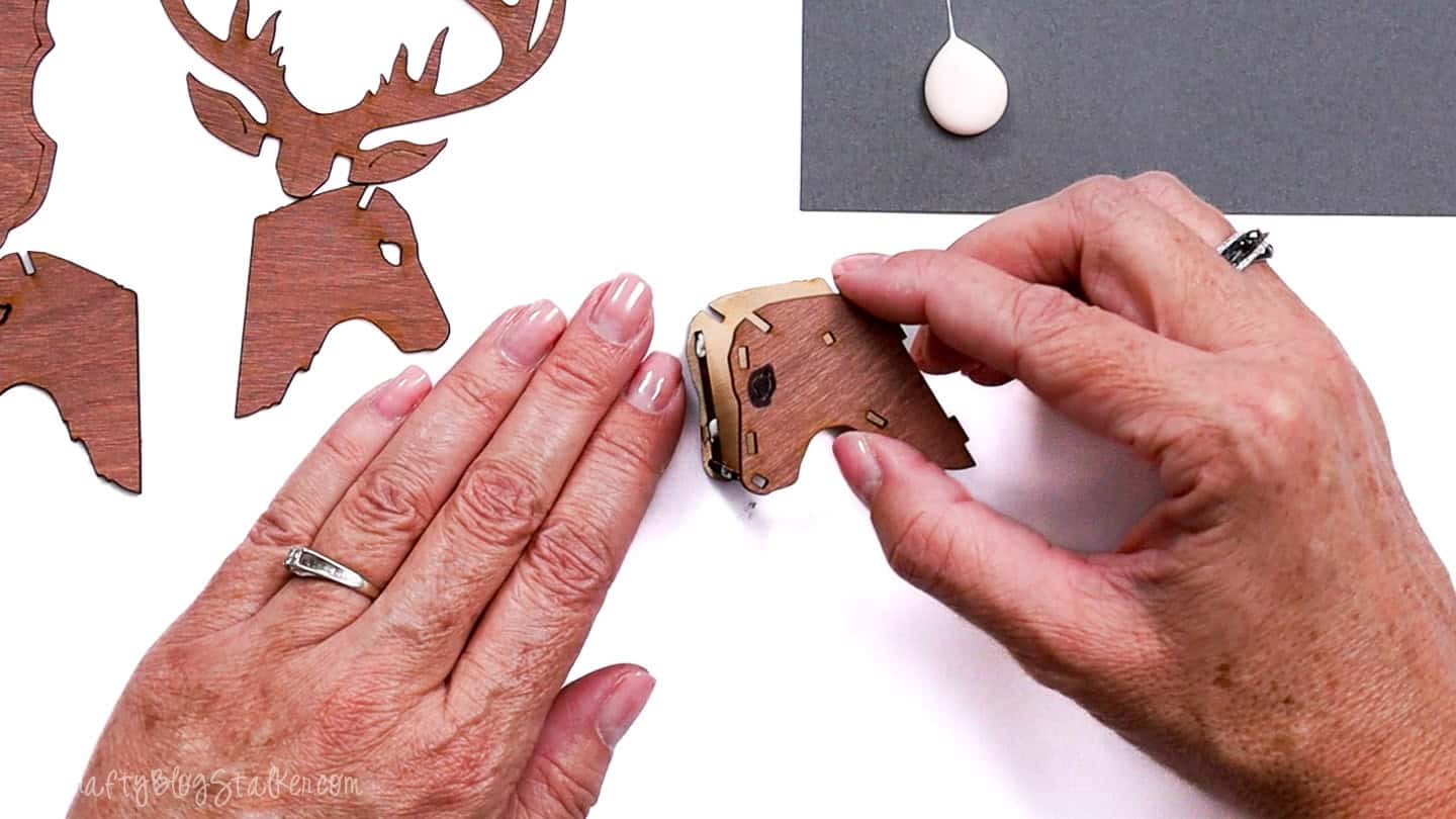 Hands assembling a small wooden reindeer head piece, with other laser-cut ornament parts and a drop of wood glue visible on the workspace.