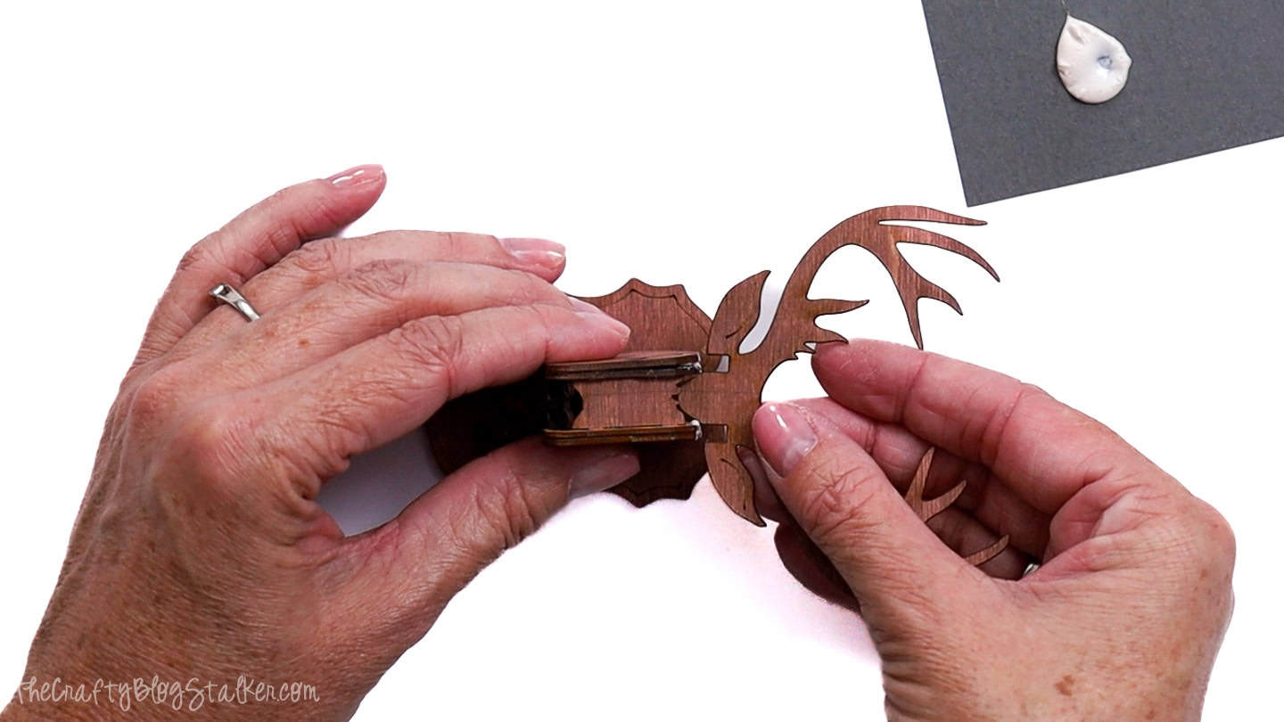 Hands attaching the wooden antler piece to the assembled reindeer head and plaque base, with wood glue visible in the corner of the workspace.