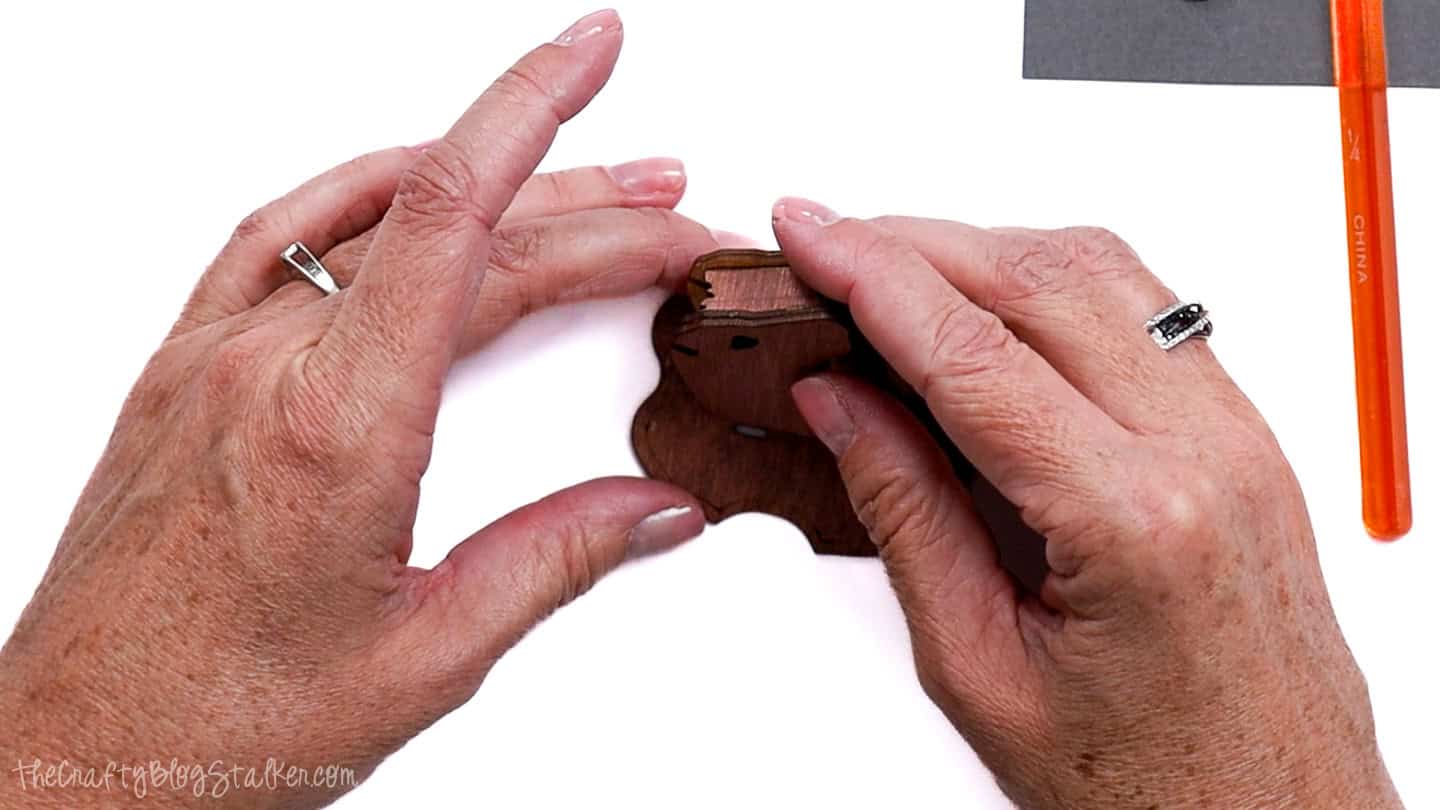Hands assembling a small wooden reindeer ornament by attaching the head piece to the base, with glue and a brush nearby.