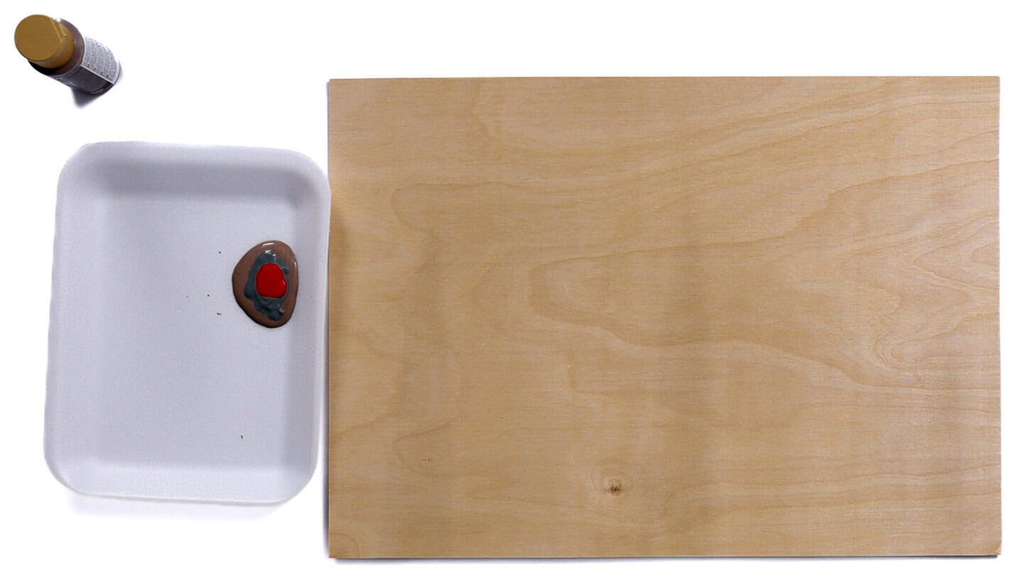 A wood board sits next to a foam tray holding small blobs of brown, gray, and red acrylic paint, with a paint bottle placed above the tray.