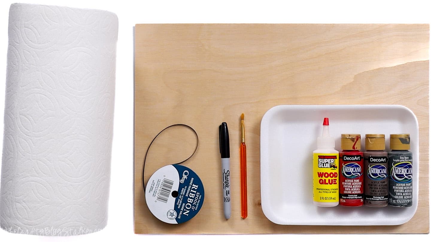 Supplies laid out for a craft project, including a roll of paper towels, a wooden board, ribbon, a marker, a paintbrush, wood glue, acrylic paints, and a foam tray.