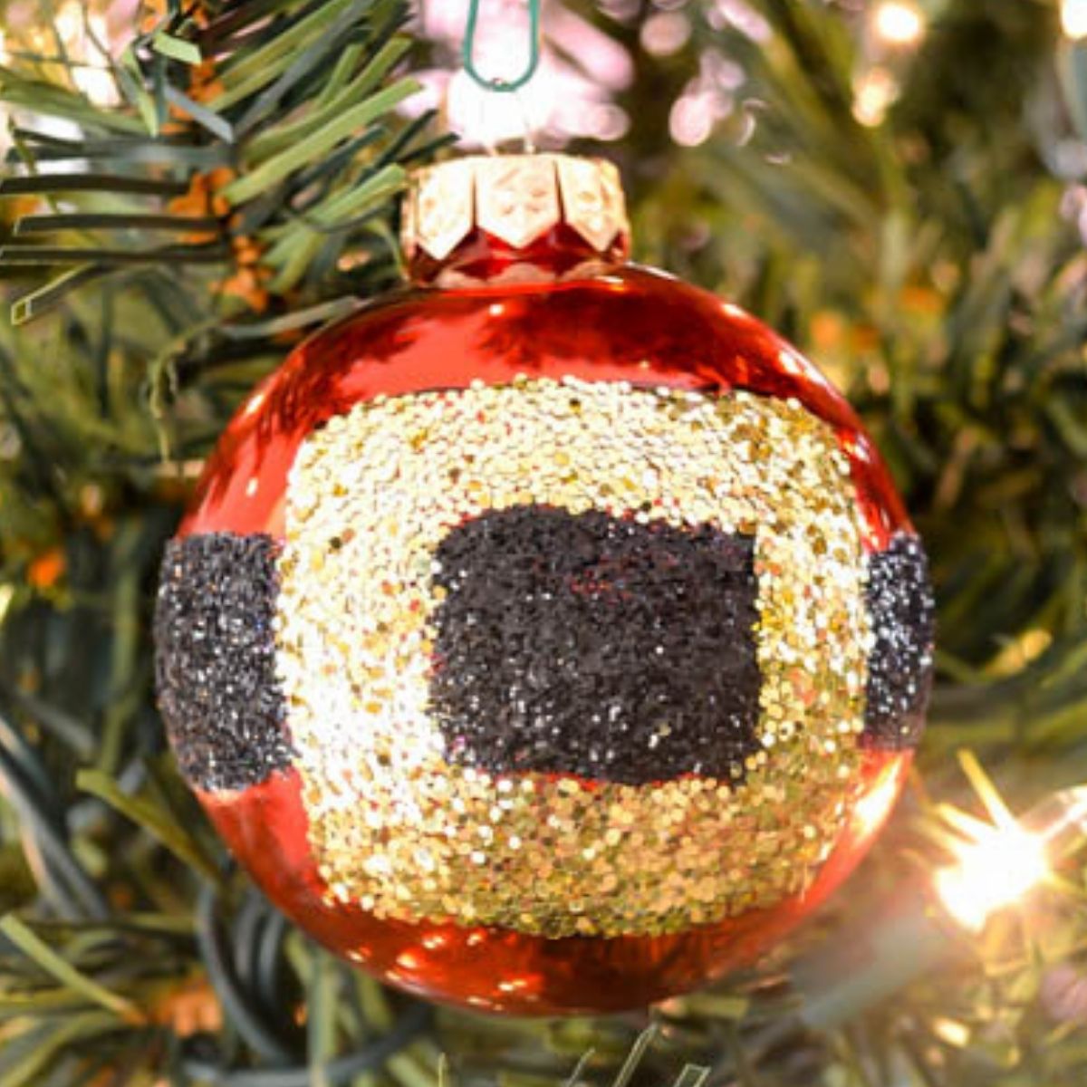 Red Christmas ornament decorated with a glittery gold belt and black buckle design, hanging on a Christmas tree branch.