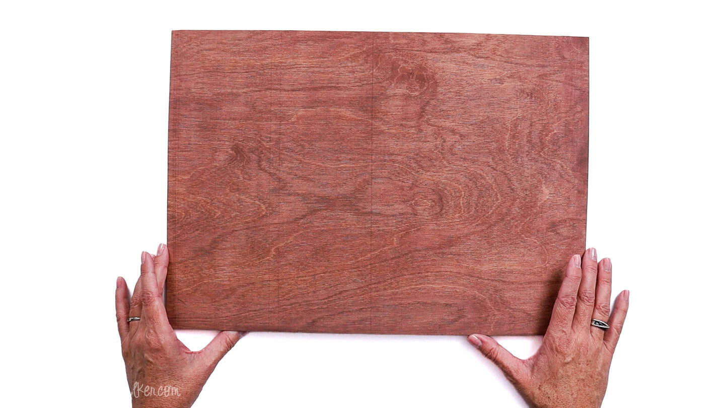 Finished stained wood board held by two hands, showing an even brown finish and visible wood grain after applying and wiping acrylic paint.