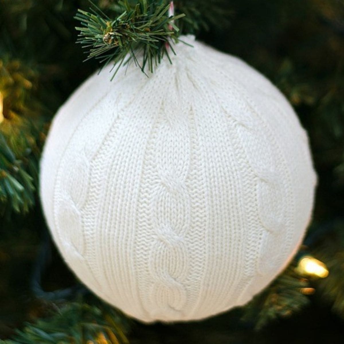 A white cable-knit sweater ornament hanging on a Christmas tree branch with green needles and soft holiday lights in the background.