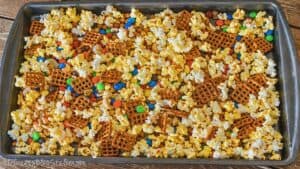 Sheet pan filled with popcorn, pretzels, nuts, and colorful candy pieces spread out before adding chocolate drizzle.
