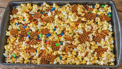 Sheet pan filled with popcorn, pretzels, nuts, and colorful candy pieces spread out before adding chocolate drizzle.