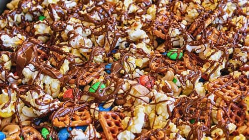 Close-up of sweet and salty snack mix with popcorn, pretzels, nuts, candy pieces, and melted chocolate drizzled on top.