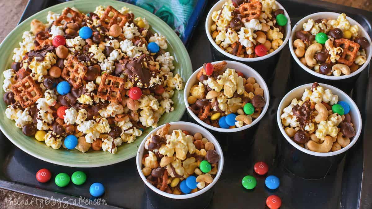 Sweet and salty snack mix with popcorn, pretzels, nuts, candy pieces, and chocolate drizzle served in a bowl and small cups.