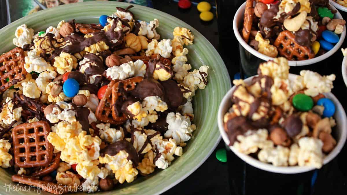 Sweet and salty snack mix with popcorn, pretzels, nuts, candy pieces, and chocolate drizzle in a bowl and small cups.