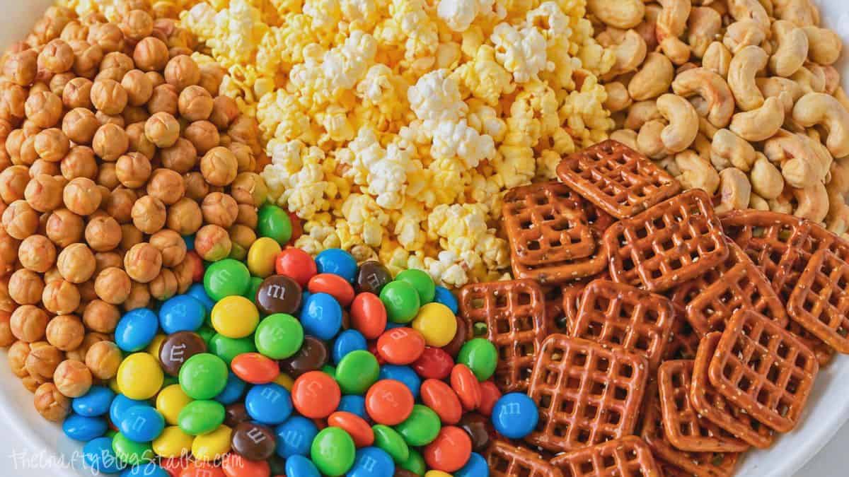 Close-up of snack mix ingredients including popcorn, pretzels, cashews, round cereal pieces, and colorful candy-coated chocolates.