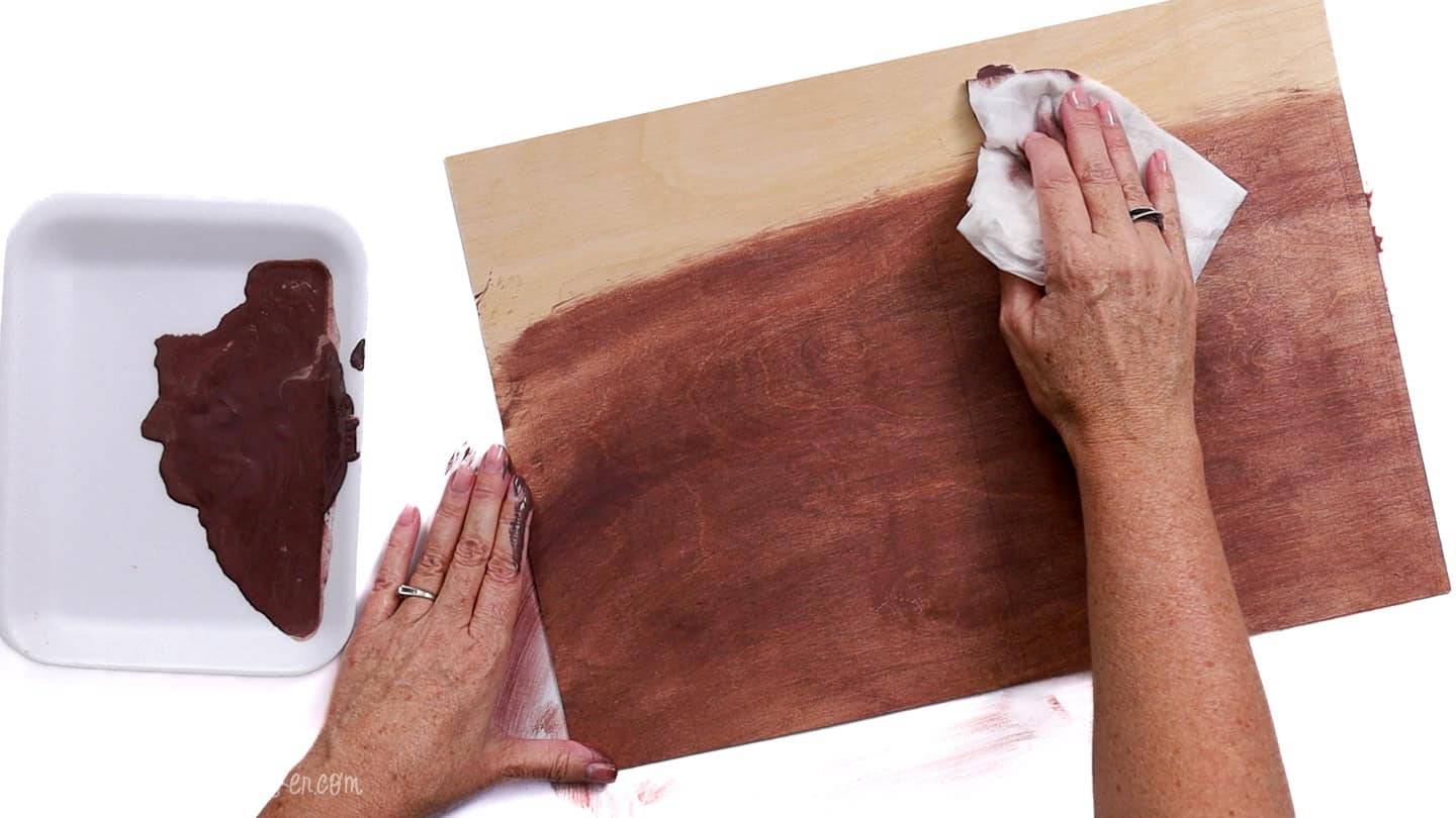 A person uses a cloth to wipe brown paint across a wood board, blending the color for a stained look. A tray of mixed paint sits nearby.