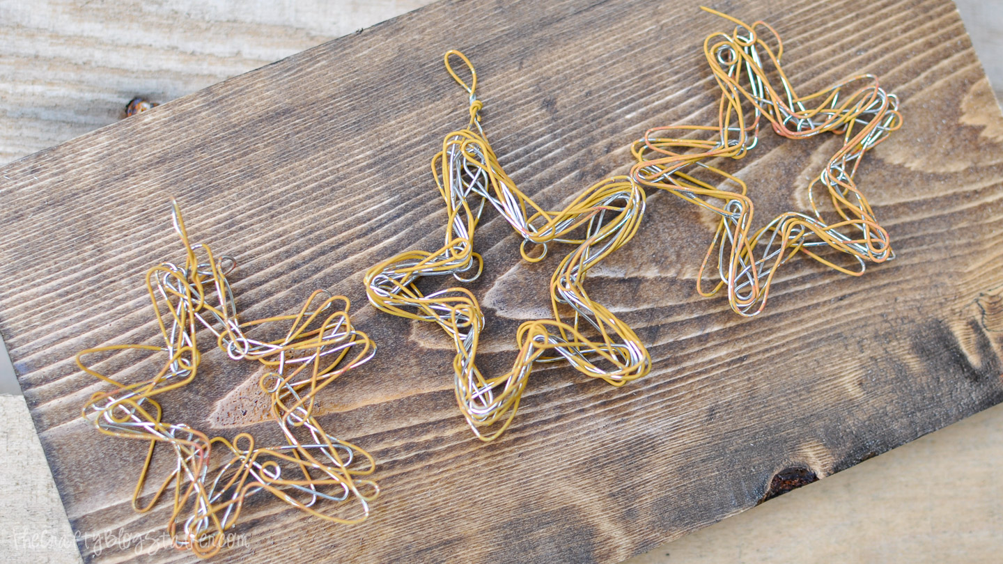 Three handmade wire star ornaments in gold and silver tones displayed on a rustic wooden board.