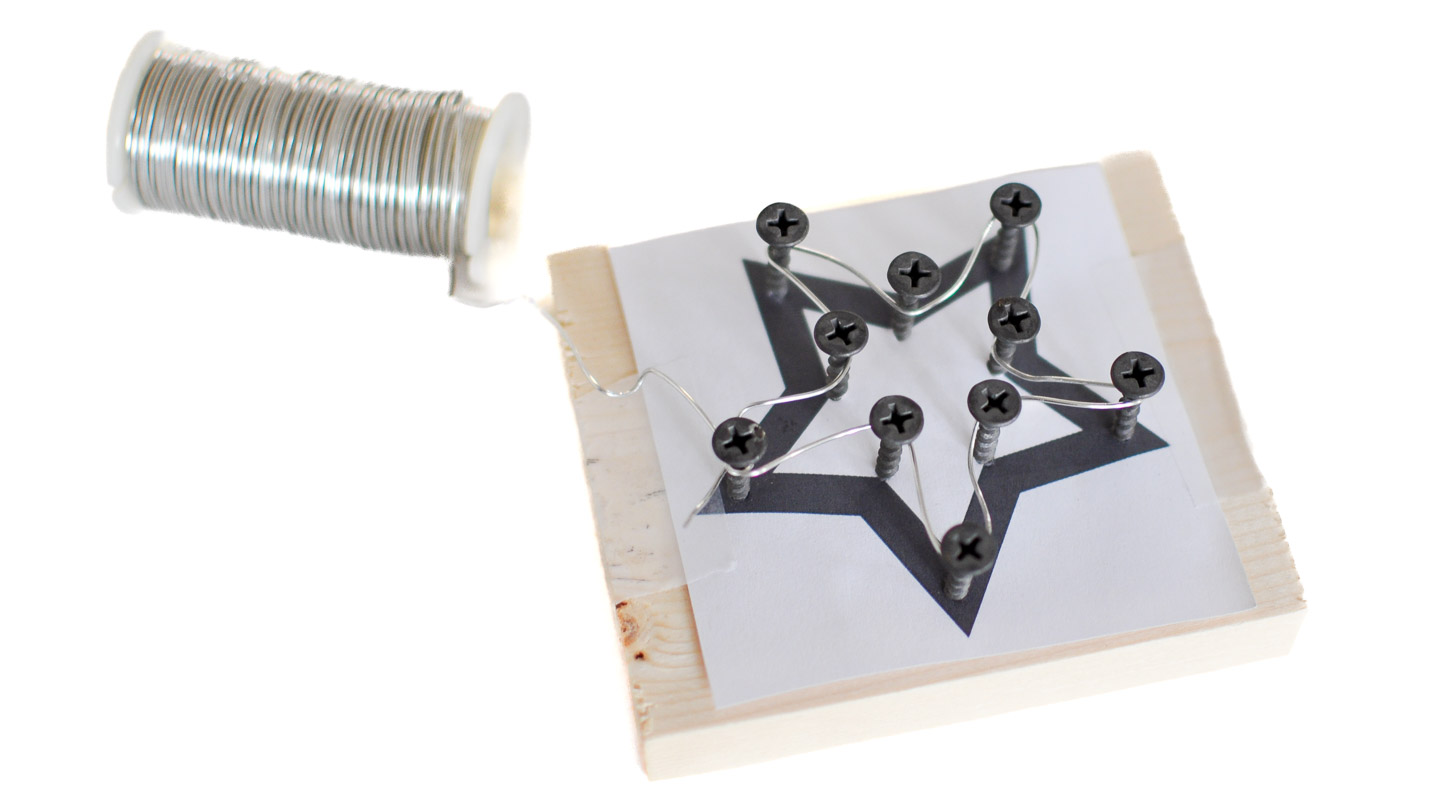 A spool of silver wire being wrapped around screws placed on a star template attached to a small wood block for a wire star ornament craft.