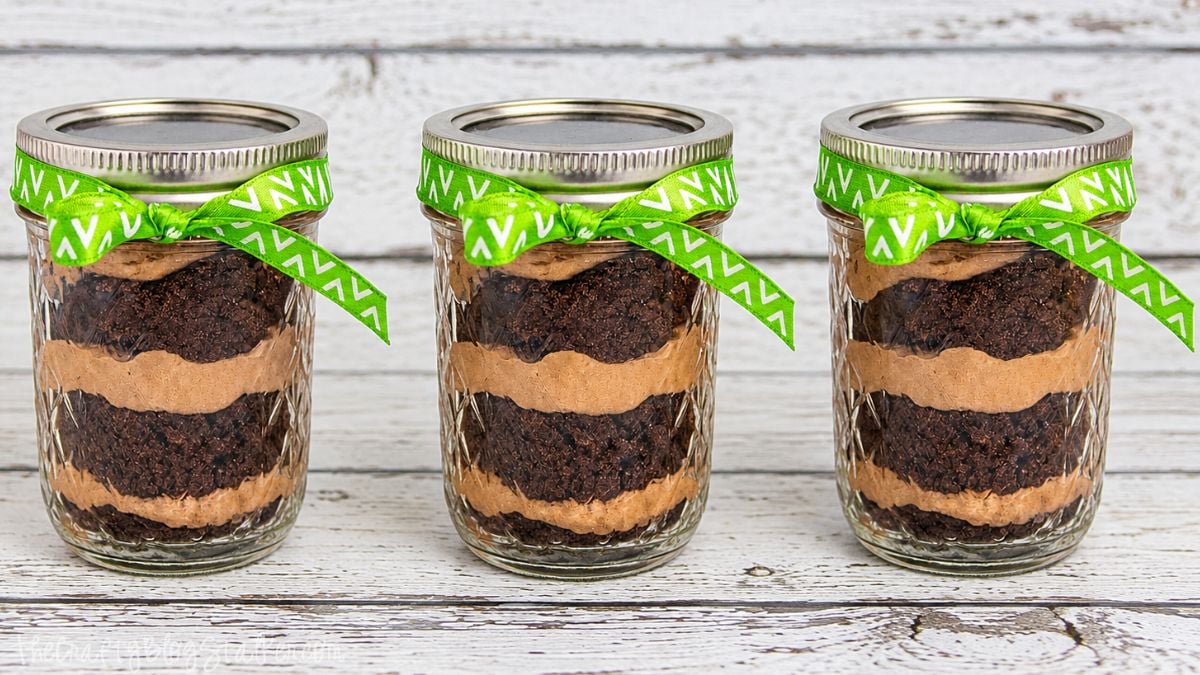 Three mason jars layered with chocolate brownie and creamy chocolate filling, each tied with a bright green ribbon on a rustic white wood surface.