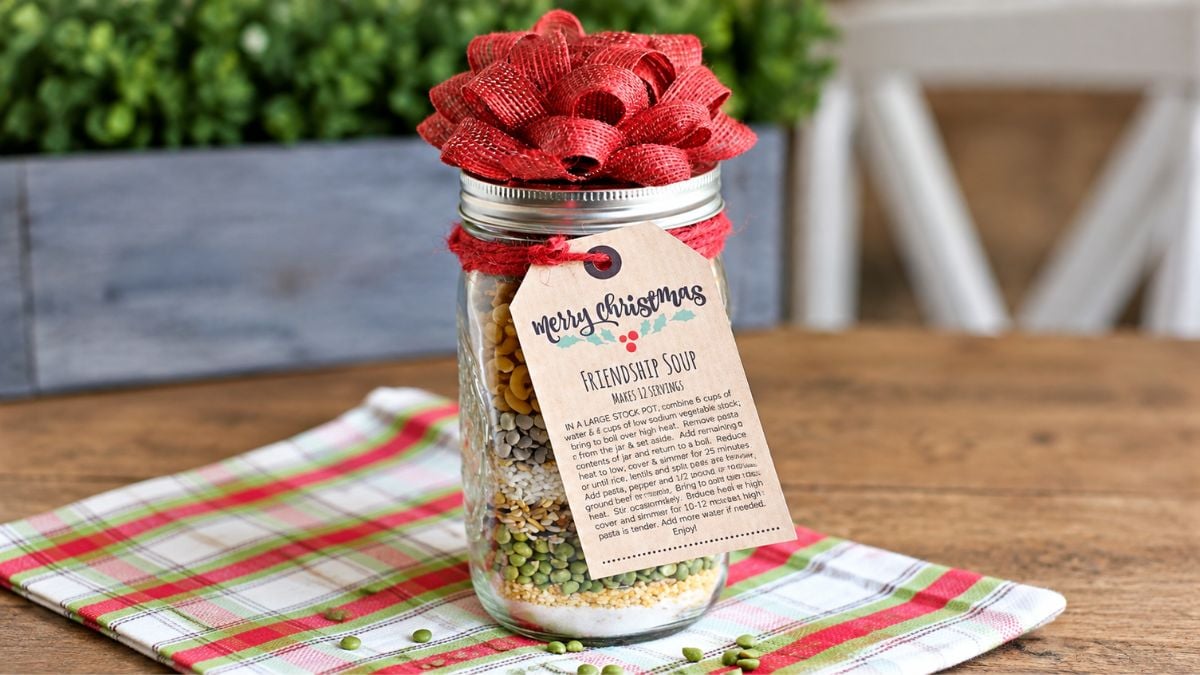 Mason jar filled with friendship soup ingredients, topped with a red bow and tied with a Merry Christmas recipe gift tag.