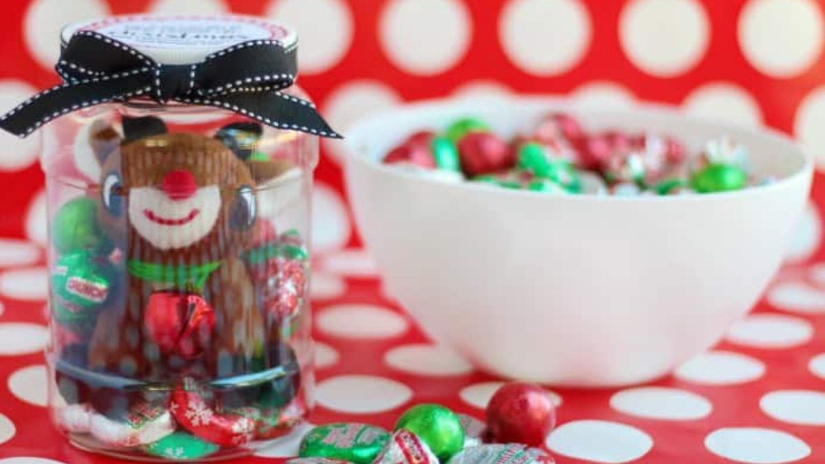Christmas countdown jar filled with chocolates and a plush reindeer, topped with a black ribbon on a red and white polka dot table.
