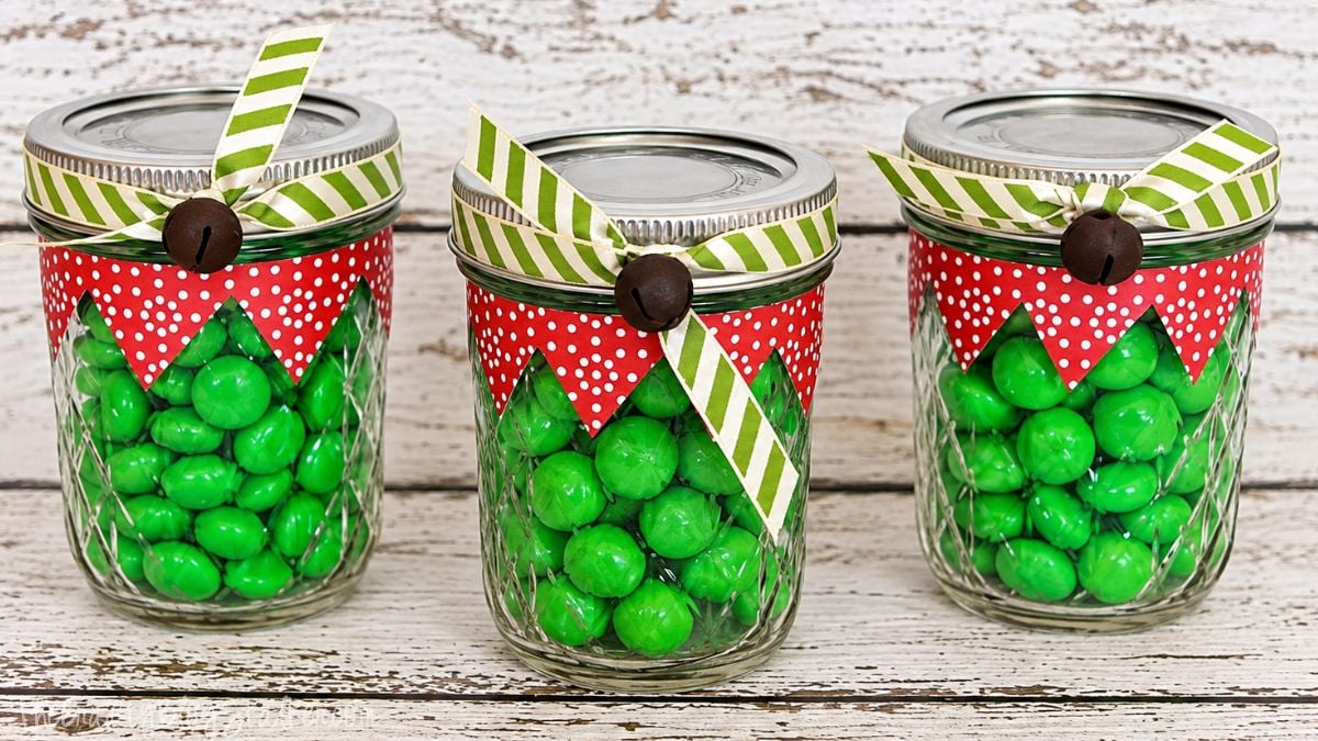 Three mason jars filled with green candy, decorated as Christmas elves with red polka-dot collars, striped ribbon, and jingle bells.