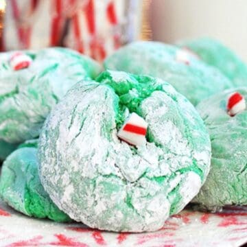 Green crinkle cookies dusted with powdered sugar and topped with small peppermint candy pieces, displayed on a festive holiday plate.