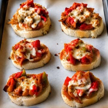 Sausage and pepper bruschetta with melted mozzarella cheese on toasted bread slices arranged on a baking sheet.