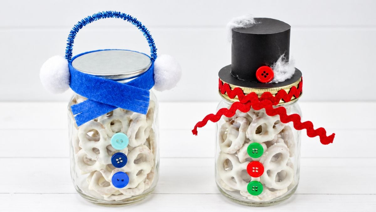 Two snowman mason jars filled with white chocolate pretzels, decorated with buttons, scarves, earmuffs, and a black top hat.