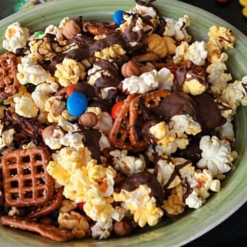 Close-up of sweet and salty snack mix with popcorn, pretzels, nuts, candy pieces, and chocolate drizzle in a bowl.