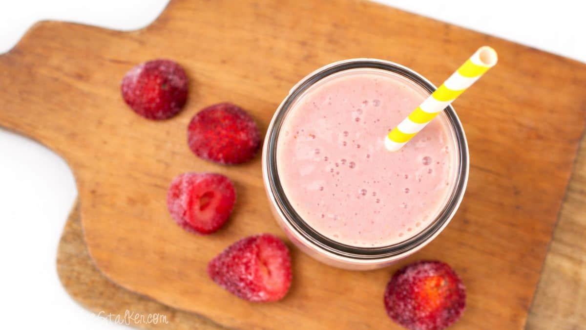 Overhead view of a strawberry smoothie in a mason jar with a striped paper straw, set on a wooden cutting board with frozen strawberries around it.