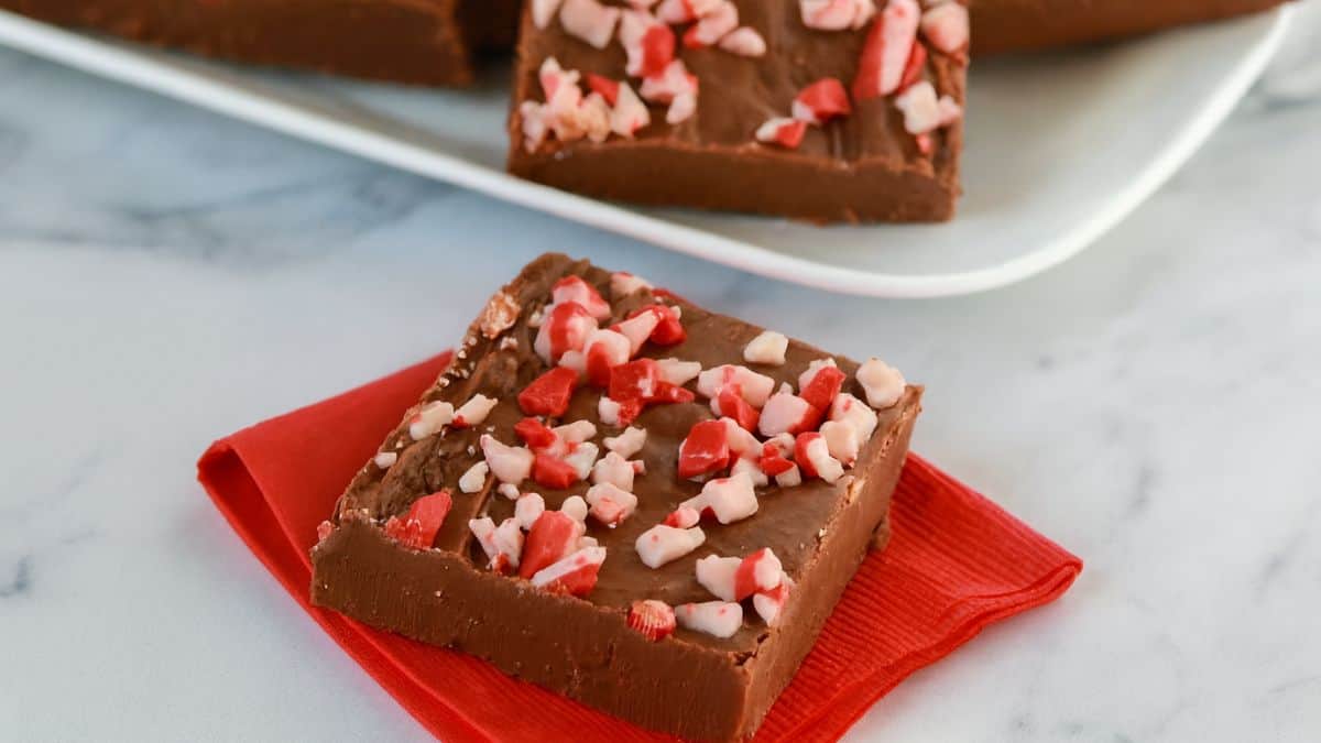 Square piece of chocolate peppermint fudge topped with crushed candy canes, served on a red napkin with more fudge pieces on a white plate in the background.
