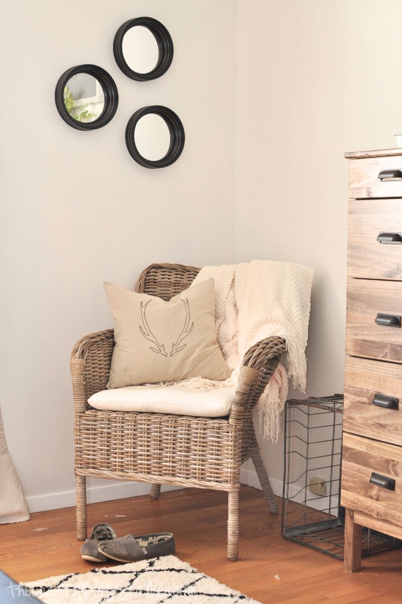 Alt text: A cozy bedroom corner with a wicker chair, antler-print pillow, soft throw blanket, black-framed mirrors on the wall, and a wooden dresser beside it.