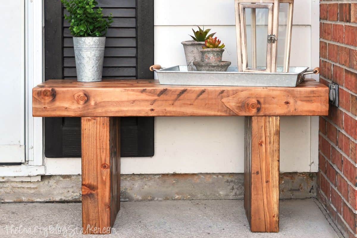 Rustic wooden outdoor bench with decor, including a metal pail with greenery, a tray of succulents, and a lantern, placed on a front porch.