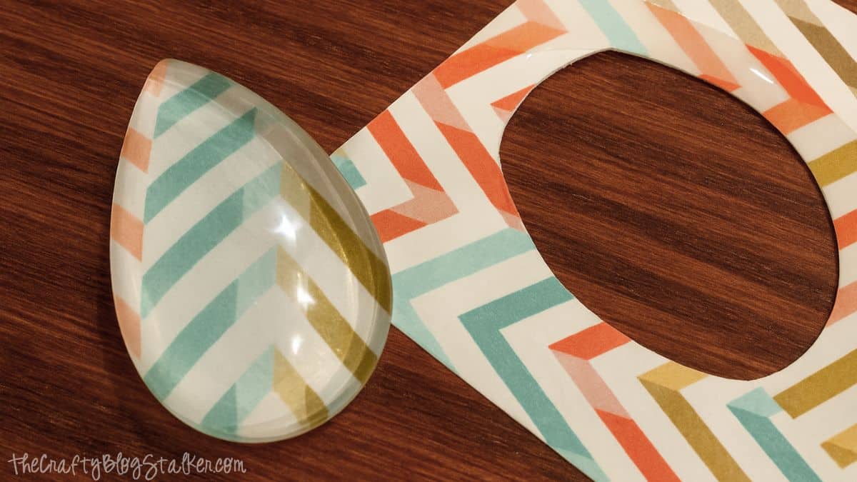 A teardrop glass cabochon sits on colorful chevron paper with a matching cutout, showing the design ready to be used for a DIY pendant necklace.