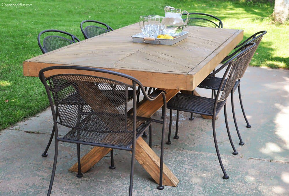 Wooden outdoor dining table with a chevron-patterned top and black metal chairs on a patio, set with a tray holding a pitcher, glasses, and lemon slices.