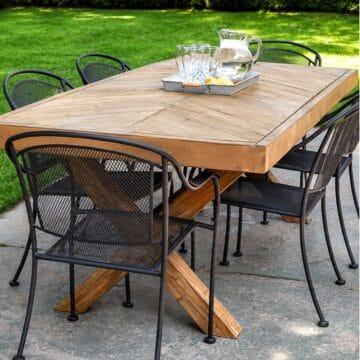 DIY outdoor farmhouse dining table with a herringbone wood top, X-shaped legs, and black metal patio chairs.