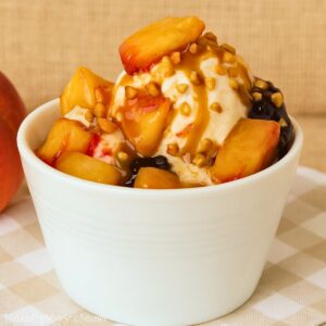 Vanilla ice cream topped with fresh peach slices, caramel sauce, chopped nuts, and hot fudge served in a white bowl.