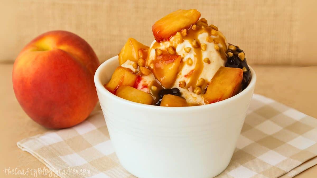 Bowl of vanilla ice cream topped with fresh peach slices, caramel sauce, and nuts, with a whole peach in the background on a checkered cloth.