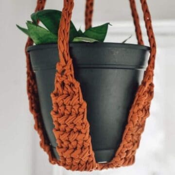 Black plant pot with green leaves hanging in a handmade rust-colored crochet plant holder made from t-shirt yarn.