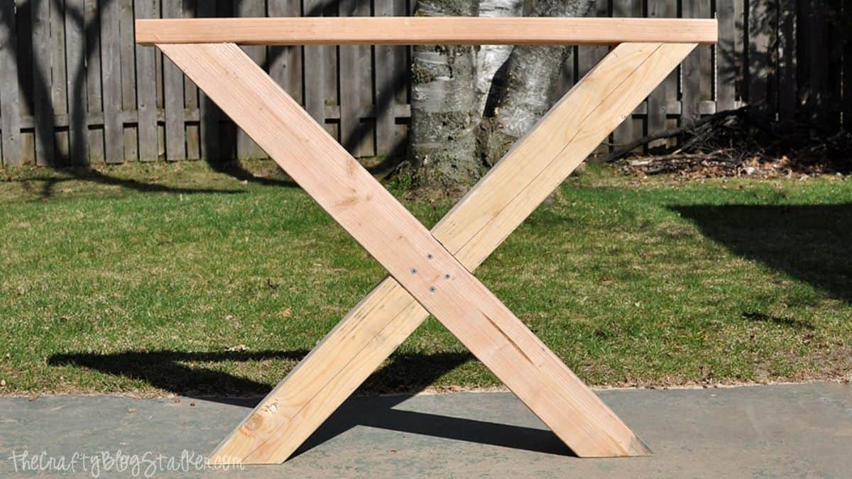 Wooden X-shaped farmhouse table leg base assembled outdoors with unfinished lumber and visible screws on a concrete patio.