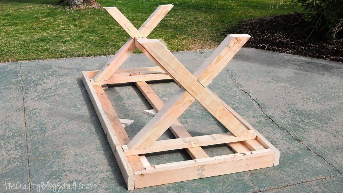 Partially assembled wooden farmhouse table base with X-shaped legs attached to the frame on an outdoor concrete patio.