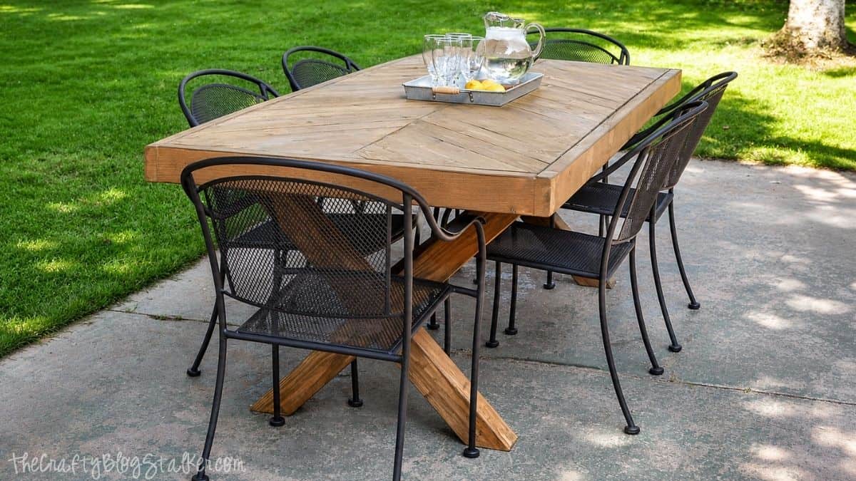 Handmade wooden outdoor dining table with a herringbone top and X-style base, surrounded by metal patio chairs on a concrete patio.