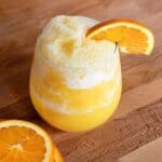 Homemade orange juice slushie in a glass with a frothy frozen texture, garnished with a fresh orange slice.