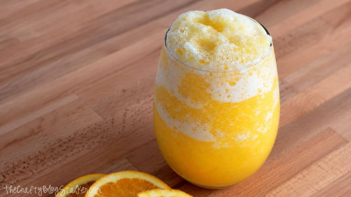Homemade orange juice slushie in a glass with a frothy frozen texture, served on a wooden surface with fresh orange slices.
