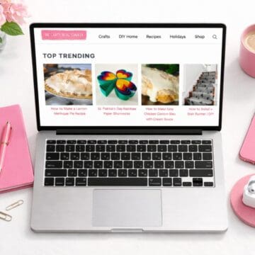 Top view of a laptop showing The Crafty Blog Stalker website on a white desk with pink notebook, pen, coffee mug, phone, earbuds, and flowers.