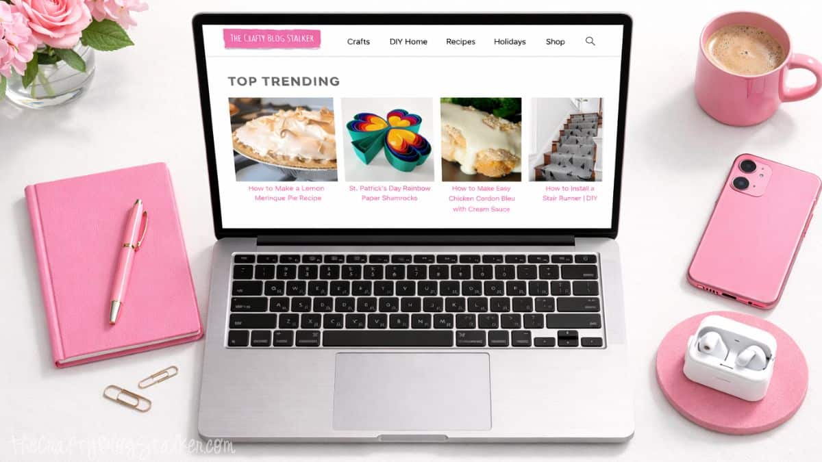 Have you been thinking of taking the plunge into blogging? With this easy step by step guide you will learn the first steps in how to start a blog! Top view of a laptop showing The Crafty Blog Stalker website on a white desk with pink notebook, pen, coffee mug, phone, earbuds, and flowers.