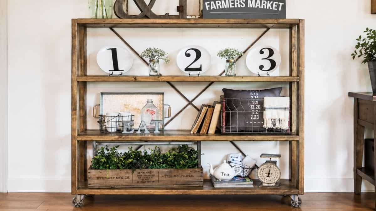 Rustic rolling bookshelf with farmhouse decor, plants, books, metal baskets, and numbered plates on wooden shelves.