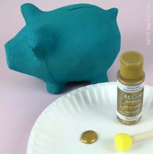gold acrylic paint and sponge dauber on a paper plate next to aa teal painted piggy bank